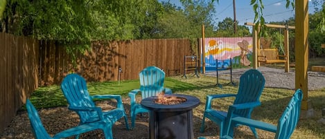 A backyard with a fire pit, four blue Adirondack chairs, and a wooden fence.