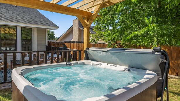 A hot tub under a wooden pergola in a backyard with a wooden fence and a house in the background. - Poolside Paradise Room Fire Pit Hot Tub (Round Rock)