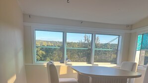 Un coin repas avec une table ronde et quatre chaises, de grandes fenêtres donnant sur la verdure et un tapis à motifs sur le sol.