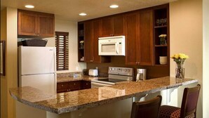 Una cocina con encimeras de granito, armarios de madera, un refrigerador, un microondas, un horno y una cafetera. Hay dos sillas y un jarrón con flores en la encimera.