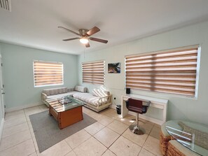 Ein Wohnzimmer mit einer Couch, einem Couchtisch und einem Sessel. Es gibt einen Deckenventilator, ein Fenster mit Jalousien und ein Bild an der Wand.