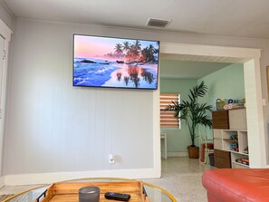 Ein Wohnzimmer mit Wand-Fernseher, der eine Strandansicht zeigt, einem Holztisch, einem roten Sofa und einem Bücherregal mit Spielzeug.