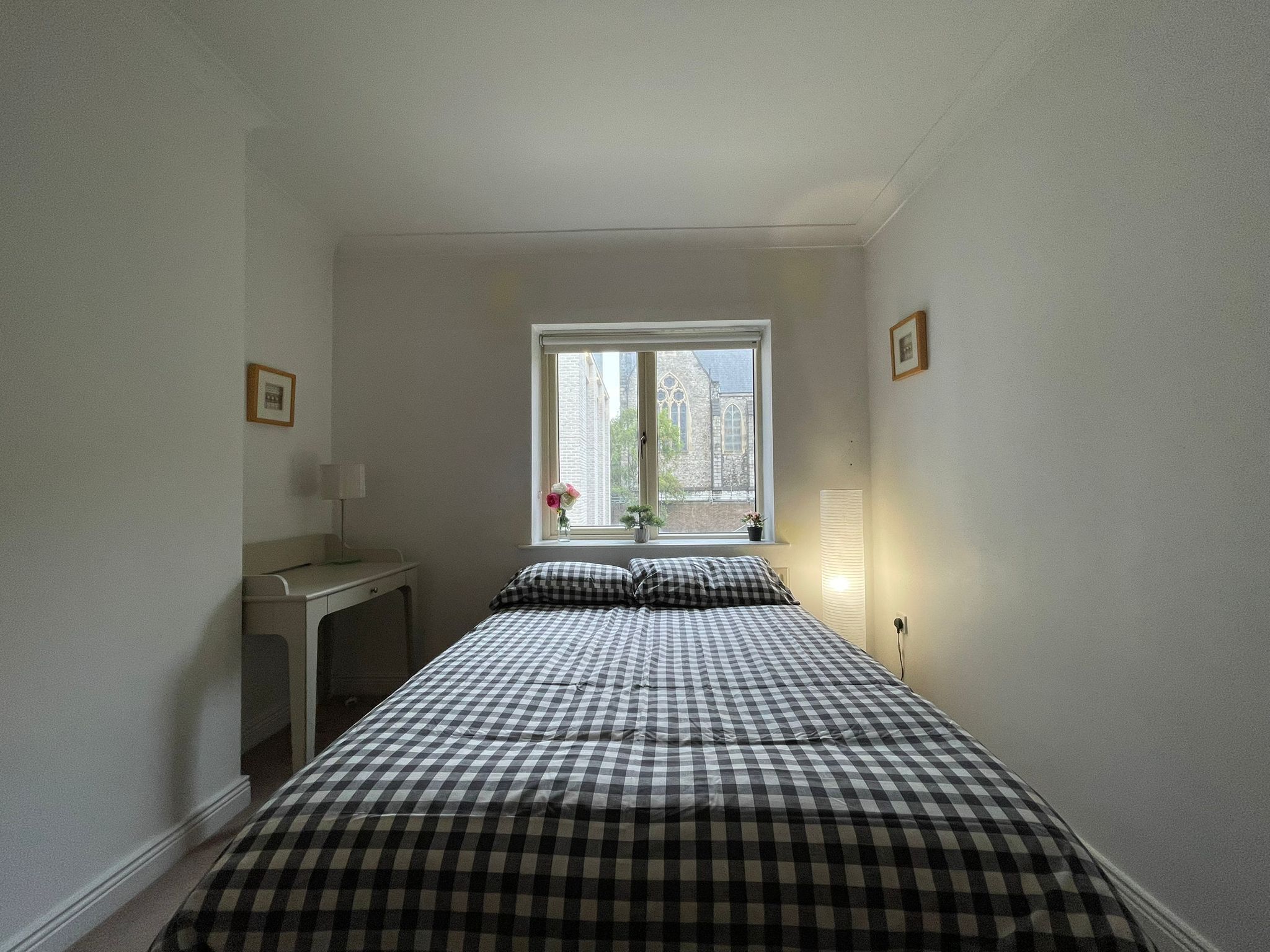 Une chambre à coucher avec une couvre-lit à carreaux, un bureau, une chaise, une fenêtre donnant sur un immeuble et une lampe de chevet.