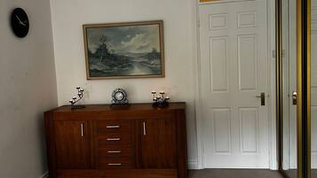 A bedroom with a checkered bedspread, a wooden dresser with a clock and candles, a painting on the wall, and a white door with a golden frame.