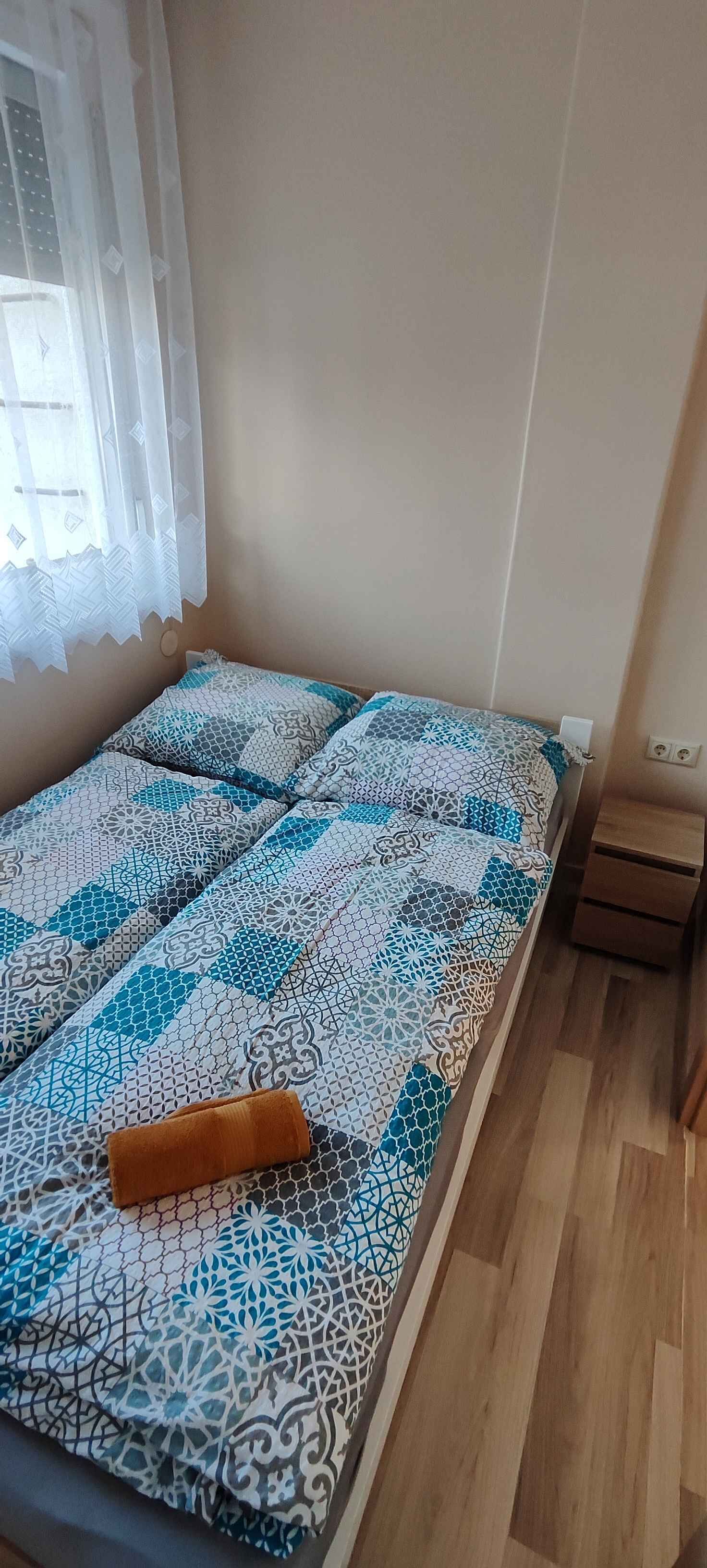 A small room with a bed covered in patterned bedding, a wooden nightstand, and a window with sheer curtains.