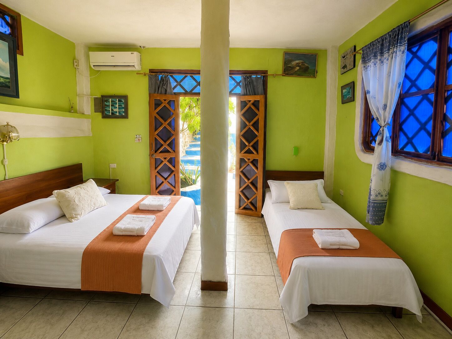 Habitación de hotel con dos camas, una ventana con cortinas azules, una puerta con celosía de madera y vistas al cielo azul y la vegetación exterior.