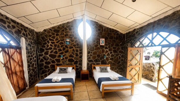 A room with two beds, stone walls, a tiled floor, and a view through a window. - PENGUIN HOSTEL (Puerto Ayora)