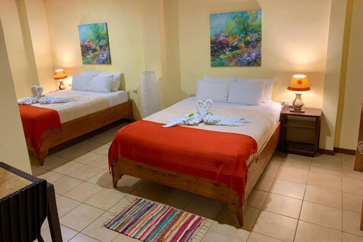 A hotel room with two beds, each with a red blanket and white pillows, and a small table with a lamp. There are two paintings on the walls and a striped rug on the floor.