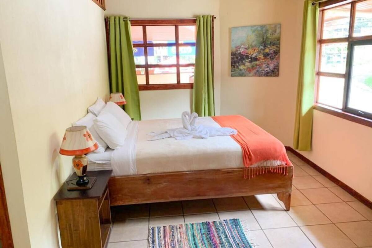 A bedroom with a bed, a nightstand, a lamp, a window with curtains, and a painting on the wall.