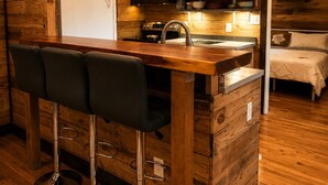 A compact kitchen with wooden walls, a central island with bar stools, a microwave, and a refrigerator. - Private Wooded Cabin w/ Hot Tub, Fire Pit. 5 minutes from Texas A&M (Navasota)