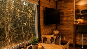 A wooden dining table with chairs, a television mounted on the wall, and a kitchen area with a countertop and cabinets. - Private Wooded Cabin w/ Hot Tub, Fire Pit. 5 minutes from Texas A&M (Navasota)