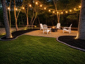 An outdoor seating area with Adirondack chairs around a fire pit, surrounded by trees and string lights. - Private Wooded Cabin w/ Hot Tub, Fire Pit. 5 minutes from Texas A&M (Navasota)