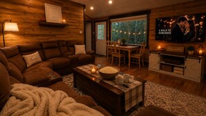 A cozy living room with a brown leather sofa, a wooden coffee table with a bowl of popcorn, a fireplace, and a television displaying a romantic scene. - Private Wooded Cabin w/ Hot Tub, Fire Pit. 5 minutes from Texas A&M (Navasota)