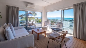 A spacious living room with a sofa, chairs, and a coffee table. Large windows offer a view of the outdoors. - Ocean View with Kiwi Vibe your perfect beach escape (Nelson)