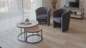 Ein modernes Wohnzimmer mit einem runden Tisch, zwei Sesseln, einem Fernseher auf einem Holzschrank und einem Balkon mit Aussicht.