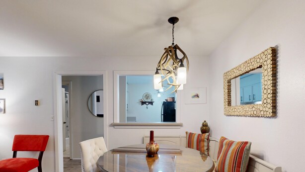 Een eetgedeelte met een ronde glazen tafel, witte gestoffeerde stoelen, een rode stoel, een kroonluchter en een spiegel met een decoratieve lijst.