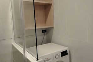 A laundry room with a washing machine, a shelf above it, and a wall-mounted electrical outlet. - View Talay Studio (Pattaya)