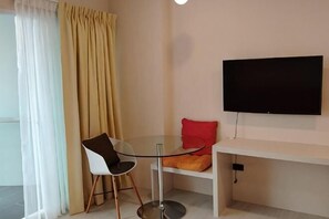 Una habitación con una mesa redonda de vidrio, una silla blanca, un cojín rojo, un banco blanco y un televisor montado.