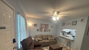 Un salon comprenant un canapé, un fauteuil et un pouf, un ventilateur de plafond, et un coin cuisine visible à travers une porte ouverte.