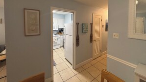 Un couloir mène à une cuisine équipée de meubles blancs, d’un réfrigérateur et d’un four à micro-ondes. Des tableaux encadrés ornent le mur, et une porte agrémentée d’un panneau décoratif est visible.
