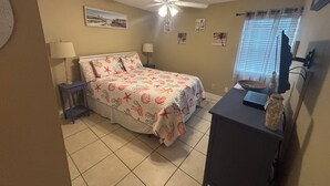 Une chambre à coucher comprenant un lit, deux tables de chevet, un bureau, un ventilateur de plafond et une fenêtre avec des stores.