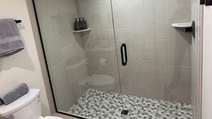 Een douche met glazen wanden en een vloer van hexagontegels, een toilet met een handdoek erop, en een plank met toiletartikelen.