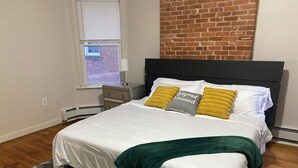 Un dormitorio con cama, mesita de noche, ventana y pared de ladrillo.