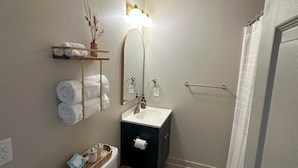 A compact bathroom with a toilet, sink, and mirror. There is a towel rack with folded towels and a small shelf with decorative items. - Cozy Studio Retreat W/ Pool Near Charlotte Airport (Gastonia)