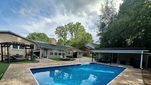 A rectangular swimming pool with a stone patio, a covered patio area, and a house with a covered garage. - Cozy Studio Retreat W/ Pool Near Charlotte Airport (Gastonia)