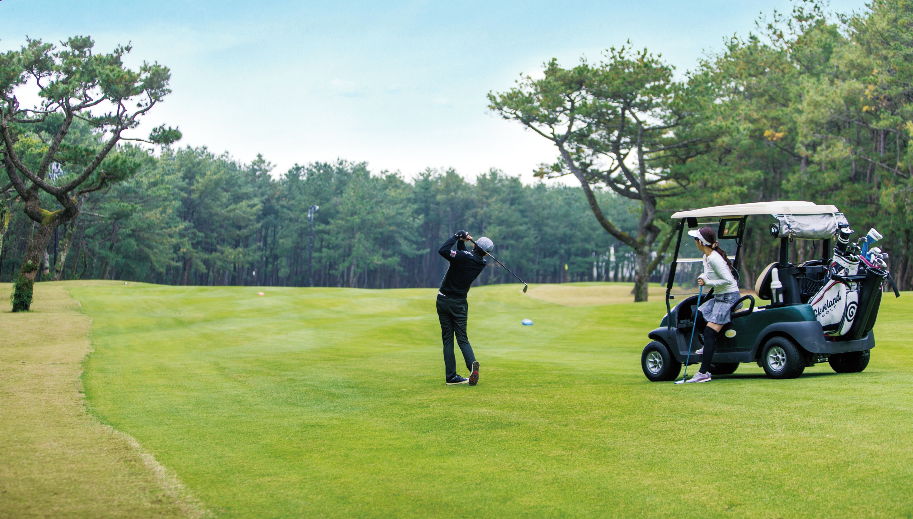 A golf course with a golfer taking a swing and a golf cart with a bag.