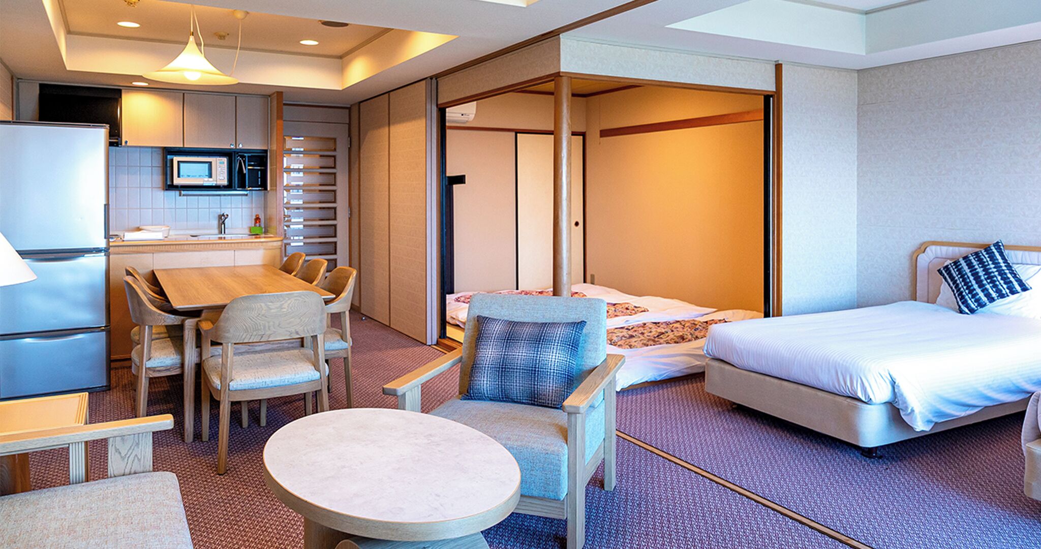 Una habitación de hotel con cama, un comedor con mesa y sillas, una pequeña cocina y un  fusible corredizo que da a otra habitación.