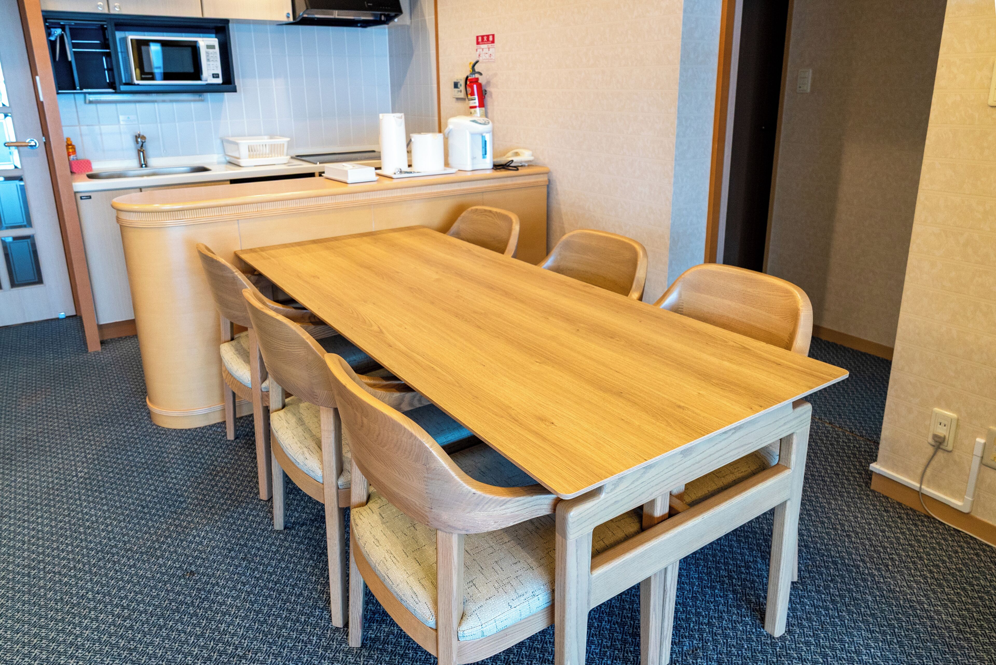 Habitación de hotel con una mesa de comedor de madera, cuatro sillas, un microondas, un lavamanos y un extintor.