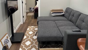 Una sala moderna con un sofá seccional gris, un televisor de pantalla plana montado en un mueble bajo, una alfombra estampada y suelo de madera.