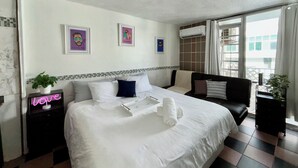 Une chambre d’hôtel avec un grand lit, une petite table de chevet surmontée d’un néon, deux tableaux encadrés accrochés au mur, un canapé noir et une fenêtre avec des rideaux.
