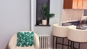 Un espacio de sala acogedor con un sillón mullido, un tapete estampado, un radiador, una ventana con planta y una zona de cocina con lavamanos y taburetes de bar.