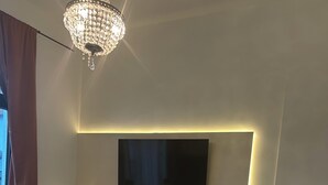Et moderne soveværelse med et vægmonteret fladskærms-tv, et sengebord med lampe, en potteplante og en rød fodskammel.