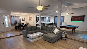 Amplia sala de estar con sofá modular, una mesa de centro y mesa de billar. Hay un comedor con candelabro y una cocina al fondo.