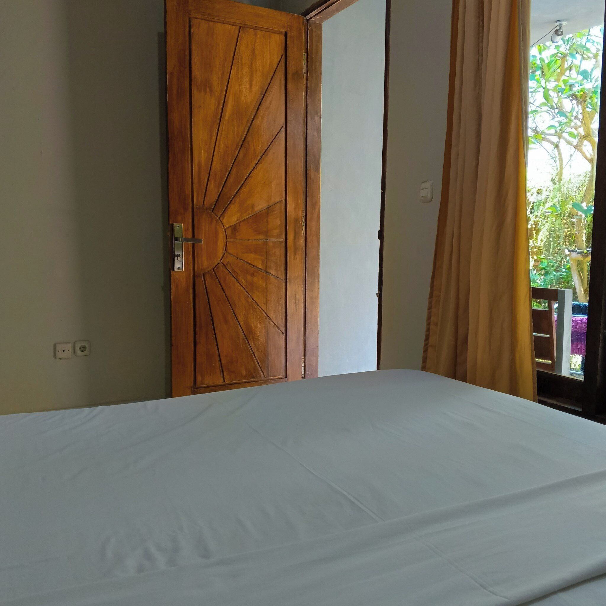 Una habitación con puerta de madera, cama con sábana blanca y vistas al exterior con vegetación.