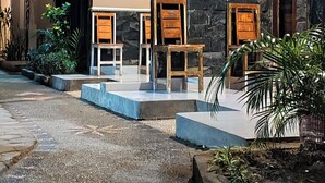 An outdoor seating area with stone walls and wooden chairs, surrounded by plants and a stone pathway. - Ayu Homestay (Kuta)