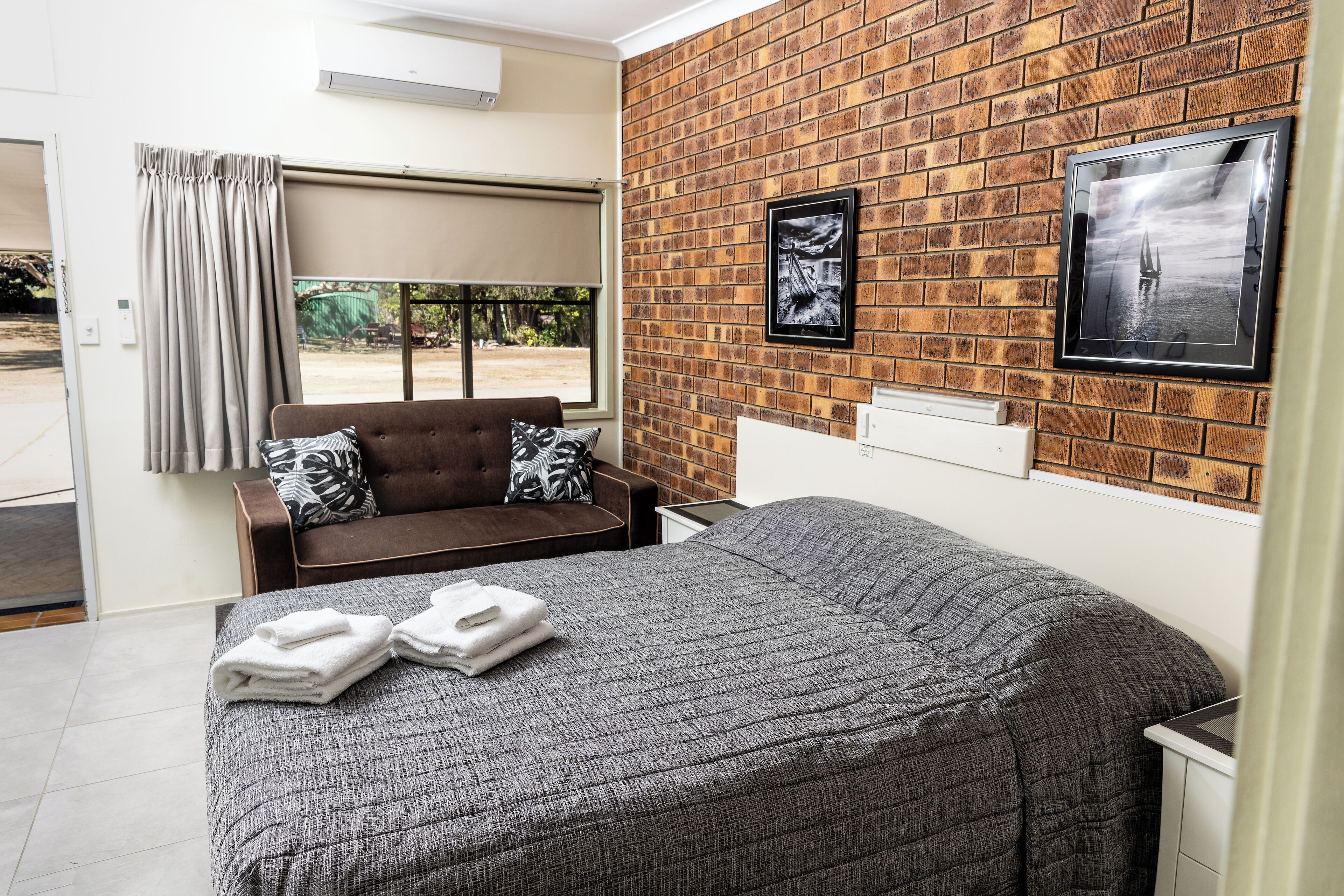 A hotel room with a bed, a brown sofa, a window with curtains, and a brick wall with framed pictures.