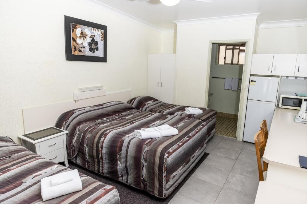 A hotel room with two beds, a small bedside table, a framed picture on the wall, a kitchenette with a refrigerator, and a door leading to a bathroom.