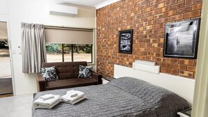 Ein Schlafzimmer mit Backsteinwand, einem Bett mit grauer Bettwäsche, einem braunen Sofa mit gemusterten Kissen und zwei gerahmten Bildern an der Wand.