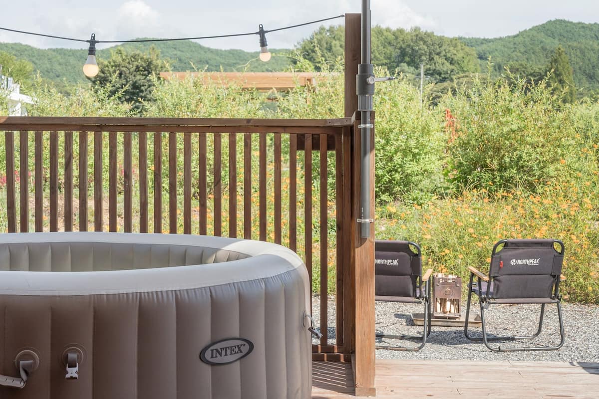 Een hottub, twee stoelen en een barbecue op een terras met een houten hek en groen in de achtergrond.
