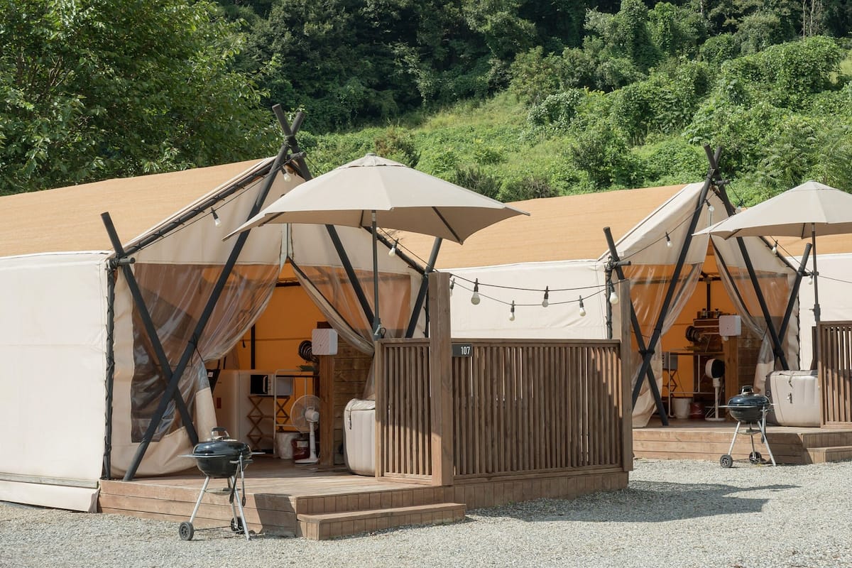 Een rij canvas tenten met luifels, elk voorzien van een barbecue en een ventilator, op een grindpad, met bomen op de achtergrond.