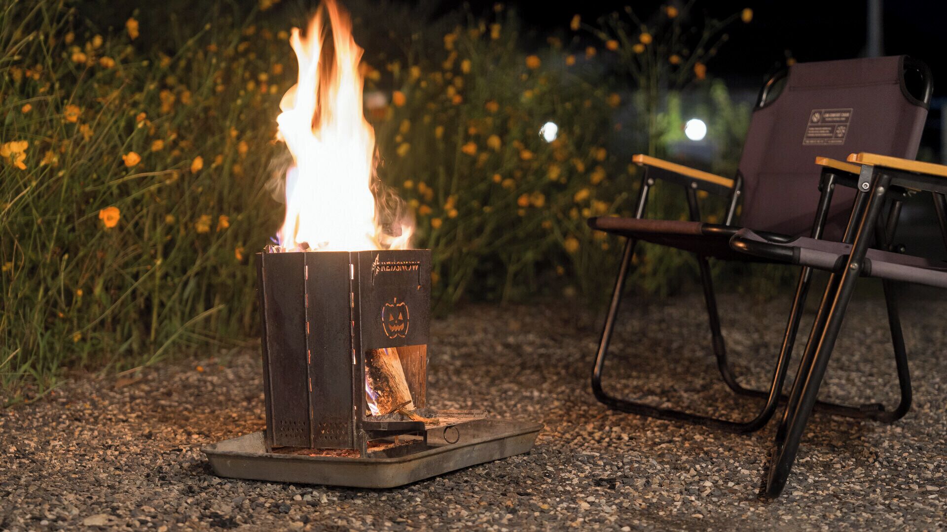 A portable fire pit with flames, surrounded by gravel, with a chair and a dark-colored bag in the background.