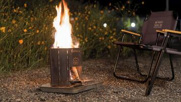 A portable fire pit with flames, surrounded by gravel, with a chair and a dark-colored bag in the background.