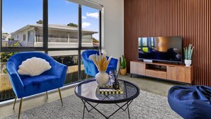 A modern living room with a large window, a TV, and blue chairs. - Riverside Modern Abode with 2 Aircons and Parking (Auckland South)