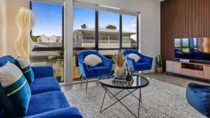 A modern living room with a blue sofa, two blue armchairs, a glass coffee table, and a flat-screen TV. - Riverside Modern Abode with 2 Aircons and Parking (Auckland South)