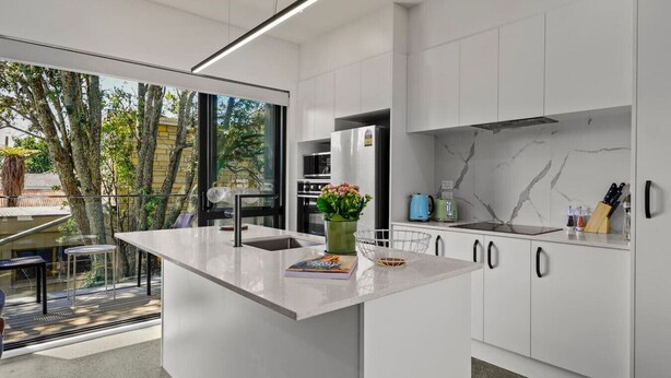 A modern kitchen with a large island, white cabinetry, and a view of trees outside. - Riverside Modern Abode with 2 Aircons and Parking (Auckland South)