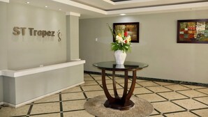 Ein Empfangsbereich mit einer Rezeption, einem an der Wand befestigten Schild mit der Aufschrift 'ST Tropez', einem runden Glastisch mit Blumenvase und zwei gerahmten Kunstwerken.